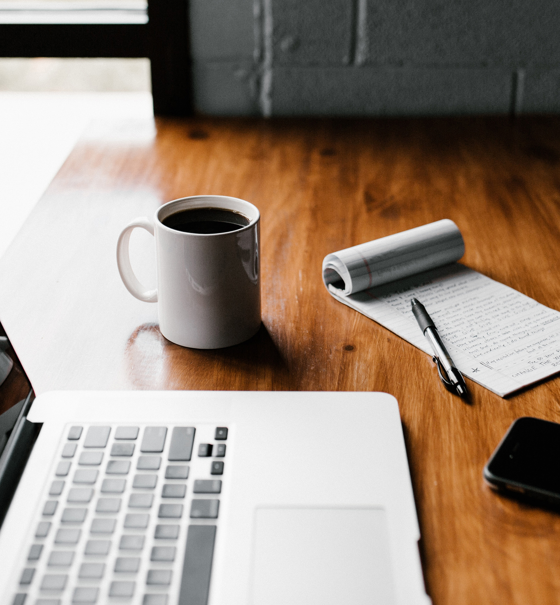 A tabletop with an open laptop, a cup of coffee, a notebook with a pen, and a cell phone.