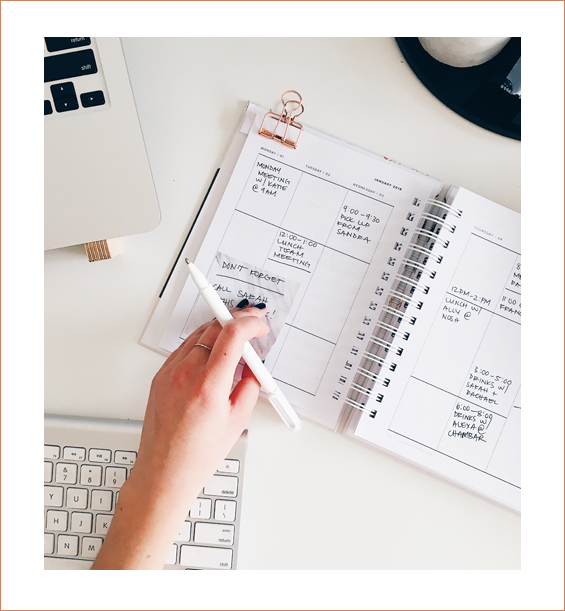 Womans hand holding a pen hovering over a planner with many things written on her schedule.