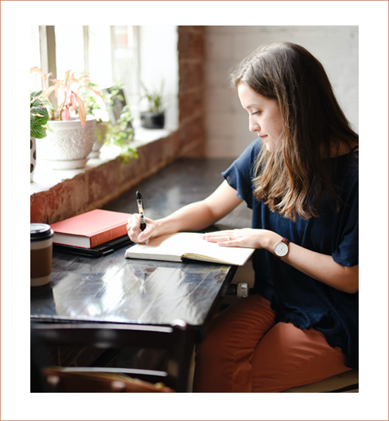 A brunette white woman sitting at a countertop writing in a notebook with books and coffee surrounding her.