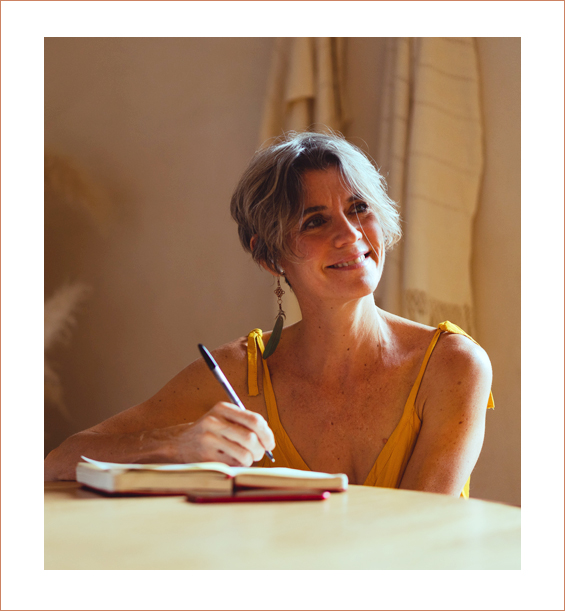 Older woman wearing a yellow sundress smiles off to the right as her pen hovers over a notebook writing.